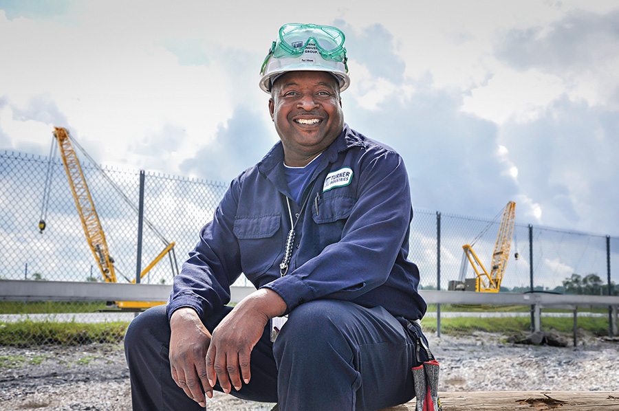 A worker in a navy jumpsuit and hard hat sits outdoors near a fence, with Project Controls cranes and cloudy skies in the background.
